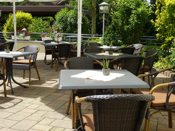 Gemütliche Café-Terrasse mit Tischen, Stühlen und üppiger grüner Bepflanzung im Hintergrund.