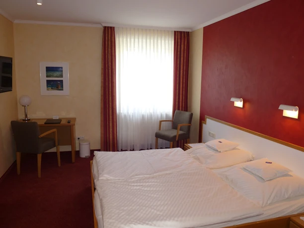 Ein gemütliches Hotelzimmer mit Doppelbett, roter Wand, Schreibtisch, Stuhl und blickdichtem Fenster.