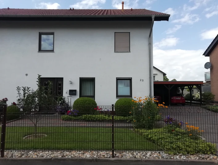 Zweistöckiges Wohnhaus mit weißer Fassade, gepflegtem Vorgarten und Carport an der rechten Seite.