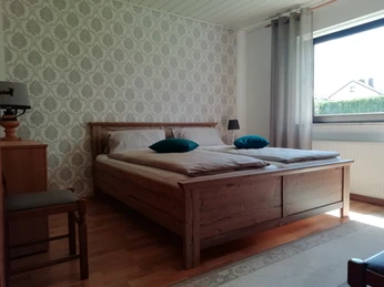 Doppelbett in hellem Raum mit Tapetenmuster, Holzmöbeln und Fenster, Tageslicht durchflutet.