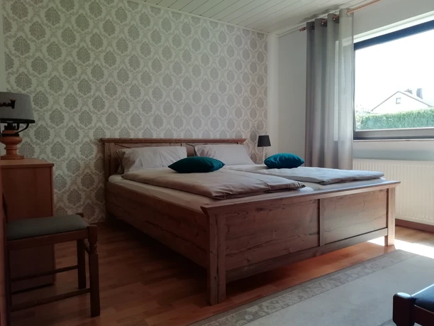 Doppelbett in hellem Raum mit Tapetenmuster, Holzmöbeln und Fenster, Tageslicht durchflutet.