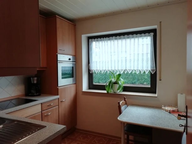 Küchenansicht 1 Helle Küche mit Holzschränken, Tisch und Stühlen am Fenster, Küchenherd und Kaffeezone sichtbar.