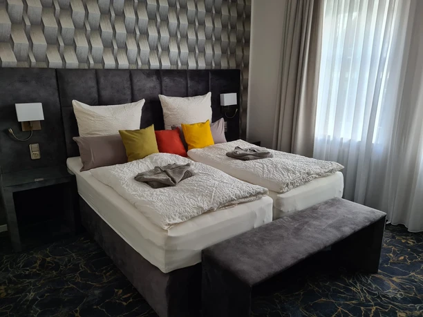 Zwei Einzelbetten mit farbigen Kissen und eleganter Tapete dahinter, Tageslicht dringt durch Vorhänge.