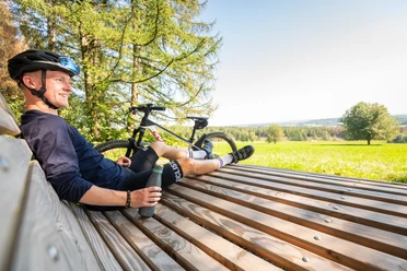 Waldsofa an der "Schönen Aussicht" bei Heckhaus Ein lächelnder Radfahrer sitzt auf einer Holzbank, genießt die Aussicht auf eine grüne Landschaft.