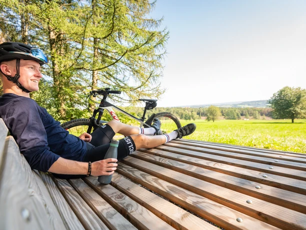 Waldsofa an der "Schönen Aussicht" bei Heckhaus Ein lächelnder Radfahrer sitzt auf einer Holzbank, genießt die Aussicht auf eine grüne Landschaft.