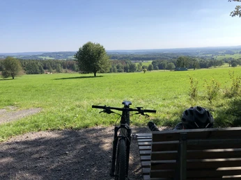 Waldsofa an der "Schönen Aussicht" bei Heckhaus Eine Parkbank mit Fahrrad im Vordergrund, bietet eine malerische Aussicht auf weitläufige grüne Wiesen.