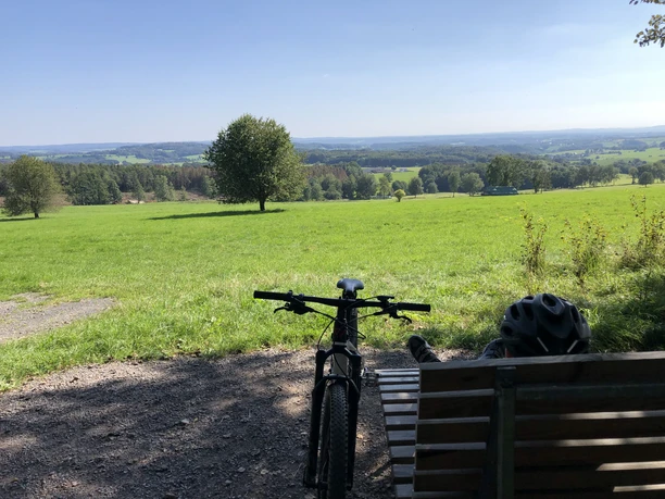 Waldsofa an der "Schönen Aussicht" bei Heckhaus Eine Parkbank mit Fahrrad im Vordergrund, bietet eine malerische Aussicht auf weitläufige grüne Wiesen.