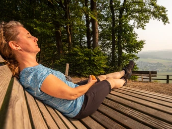 Aussichtspunkt Stachelhardt Frau entspannt auf Holzbank in hügeliger Landschaft bei sonnigem Wetter, Bäume im Hintergrund.