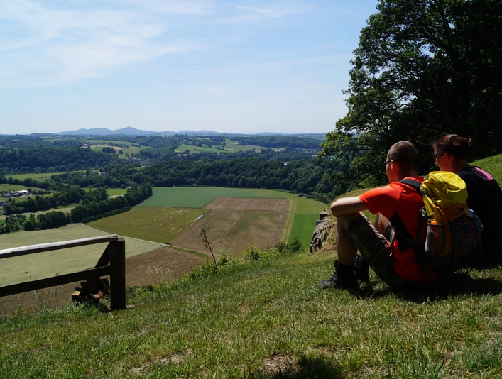 Ausblick vom Stachelberg Zwei Wanderer sitzen auf einer grünen Wiese mit Blick auf ein weites, hügeliges Tal unter blauem Himmel.