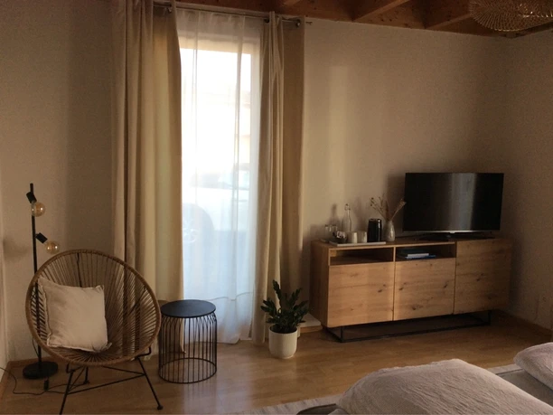 Gemütliches Wohnzimmer mit TV, Korbsessel und Lampe. Helle Vorhänge lassen Tageslicht herein.
