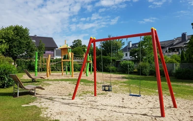 Kinderspielplatz mit Schaukeln, Klettergerüst und Rutsche in grünem Garten unter blauem Himmel.