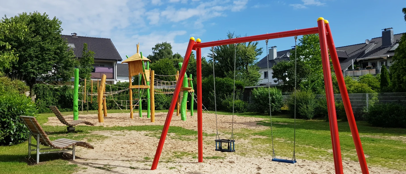 Kinderspielplatz mit Schaukeln, Klettergerüst und Rutsche in grünem Garten unter blauem Himmel.