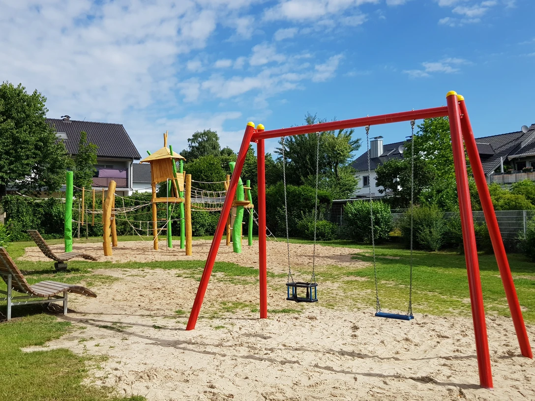Spielplatz Dahlienweg Kinderspielplatz mit Schaukeln, Klettergerüst und Rutsche in grünem Garten unter blauem Himmel.