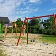 Spielplatz Dahlienweg Kinderspielplatz mit Schaukeln, Klettergerüst und Rutsche in grünem Garten unter blauem Himmel.
