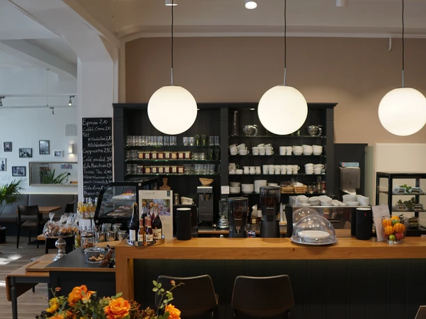 Einladende Café-Theke mit Pendelleuchten, Regalen voller Gläser und Tassen, sowie frischen Blumen.