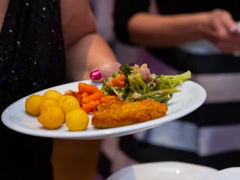 Hotel Adelmann_Speisen Eine Person hält einen Teller mit paniertem Schnitzel, Kroketten, Möhren und Broccoli. Im Hintergrund serviert jemand Salat.