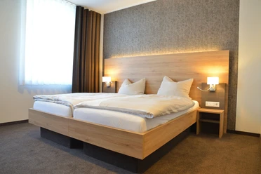 Hotel Adelmann_Zimmer Modernes Hotelzimmer mit Doppelbett, grauer Akzentwand, großen Fenster und wärmender Beleuchtung.
