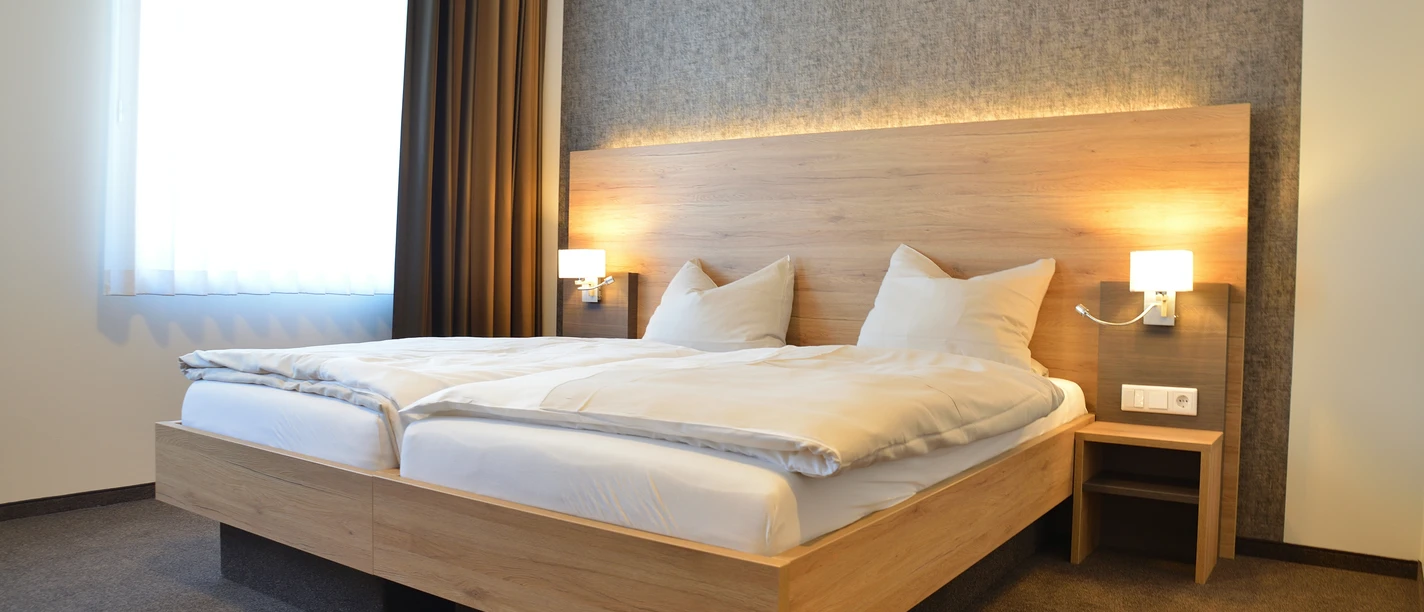 Modernes Hotelzimmer mit Doppelbett, grauer Akzentwand, großen Fenster und wärmender Beleuchtung.