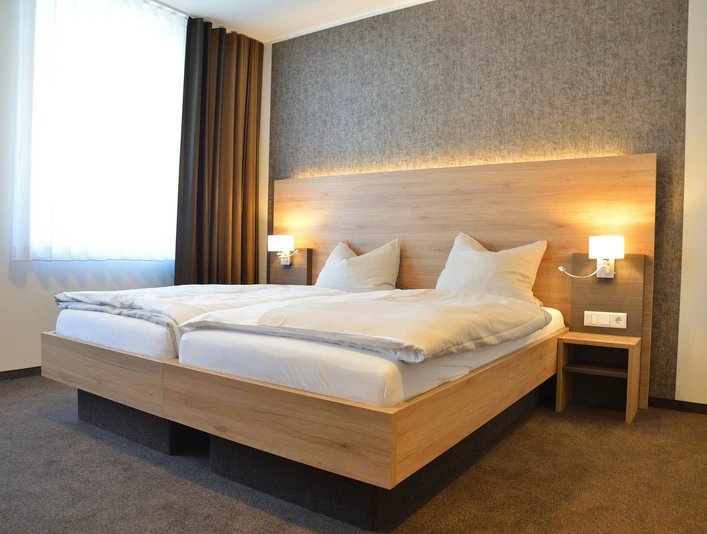 Hotel Adelmann_Zimmer Modernes Hotelzimmer mit Doppelbett, grauer Akzentwand, großen Fenster und wärmender Beleuchtung.