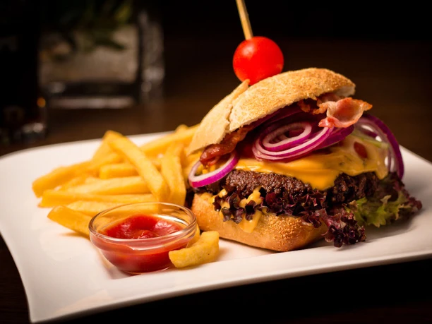 Hotel Adelmann_Speisen Ein saftiger Burger mit Käse, roten Zwiebeln und Speck, serviert mit Pommes und Ketchup auf einem Teller.