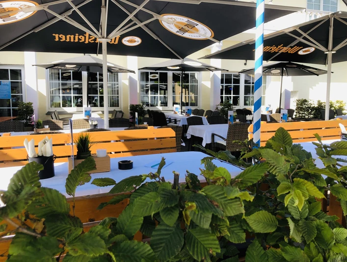 Sonniger Biergarten mit gelben Holzbänken, blühendem Grün und großen schwarzen Sonnenschirmen.