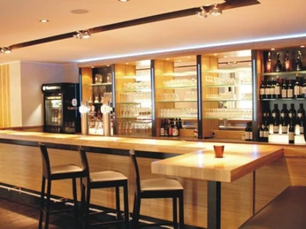 Moderne Bar mit Holztheke, eleganten Hockern und beleuchtetem Regal voller Gläser und Flaschen.