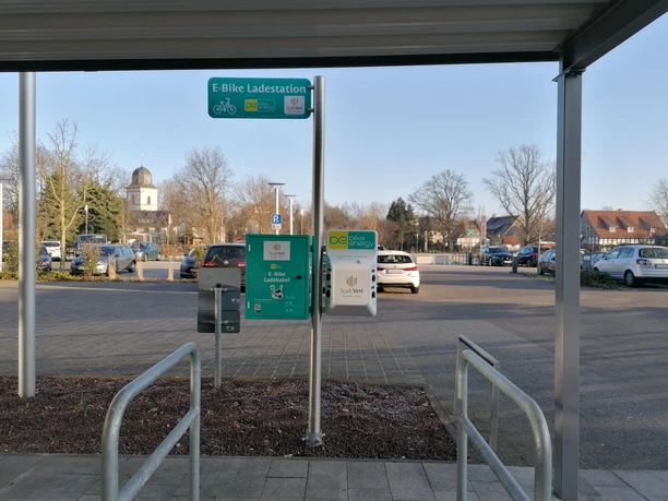 Parkplatz mit E-Bike-Ladestation, umgeben von kahlen Bäumen und einem Kirchturm im Hintergrund.