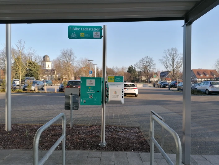 Parkplatz mit E-Bike-Ladestation, umgeben von kahlen Bäumen und einem Kirchturm im Hintergrund.