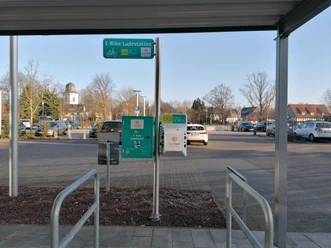 Ladestation am Rathaus mit Blick auf die Bürmsche Wiese Parkplatz mit E-Bike-Ladestation, umgeben von kahlen Bäumen und einem Kirchturm im Hintergrund.