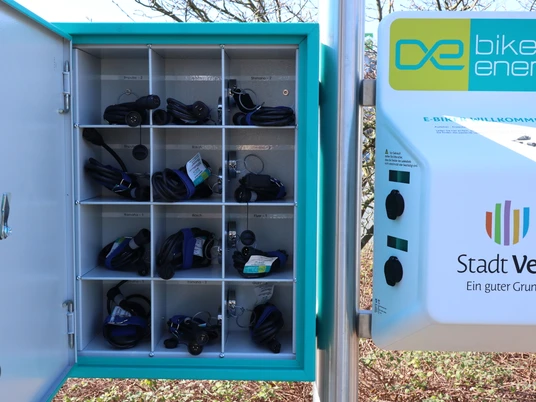 Ladestation für E-Bikes mit offenen Fächern für Ladekabel in einem grünen städtischen Umfeld.