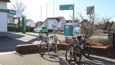 Fahrräder parken bei einer E-Bike-Ladestation an einer Straße, im Hintergrund sind Häuser zu sehen.