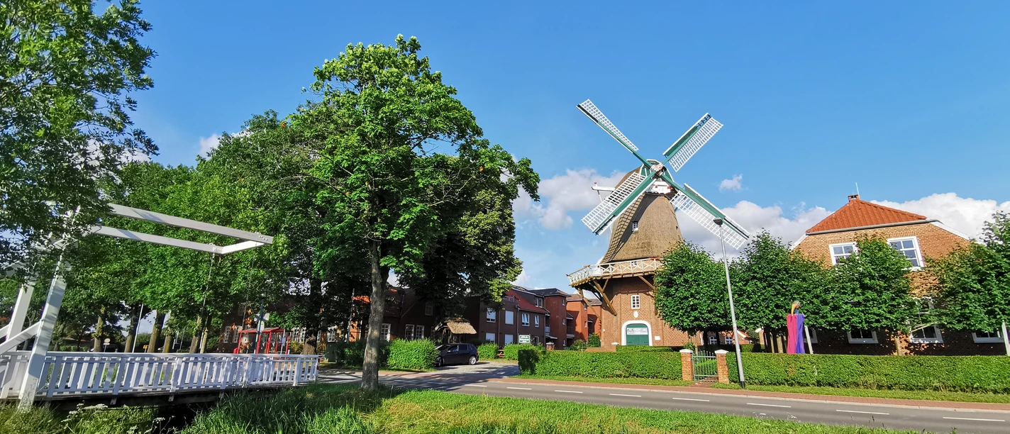 Rhauderfehn_Mühle Hahnentange_01.02.21_mk (6).jpg Historische Windmühle mit vier Flügeln neben einem Kanal, umgeben von Bäumen und Wohnhäusern unter blauem Himmel.