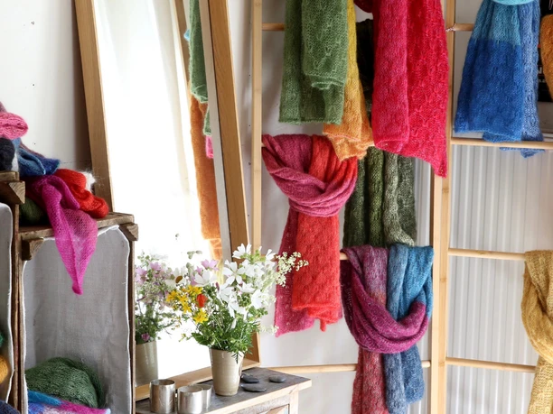 Auswahl Handgestrickte bunte Schals aus weicher Wolle hängen an Holzleitern, davor ein Wiesenblumenstrauß.