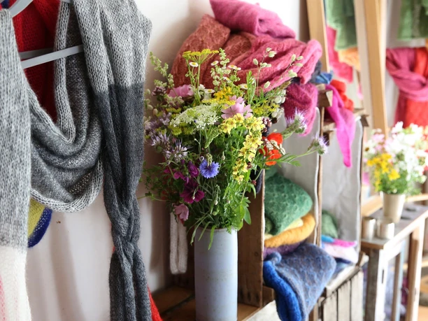 Mode Taimi Design Ein grauer Schal und eine Vase mit bunten Wiesenblumen vor Regalen voller farbiger Strickwaren.