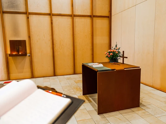 Hamm2022_Autobahnkapelle05_CCBYSA_ThorstenHübnerStadtHamm.jpg Schlichter Andachtsraum mit modernem Lesepult und Blumenschmuck auf hellem Steinboden.