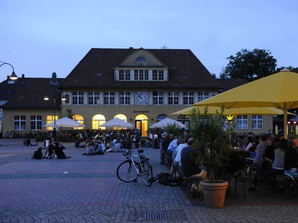 Studieren-Leben-Siggi-Bielefeld.jpg Historisches Gebäude mit beleuchtetem Zifferblatt; Menschen genießen den Abend unter gelben Sonnenschirmen.