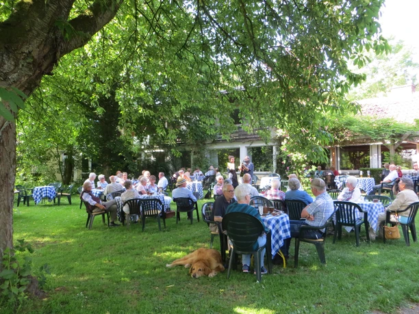 Menschen sitzen an blauen Tischen im Freien, umgeben von Bäumen, während ein Hund auf dem Rasen ruht.