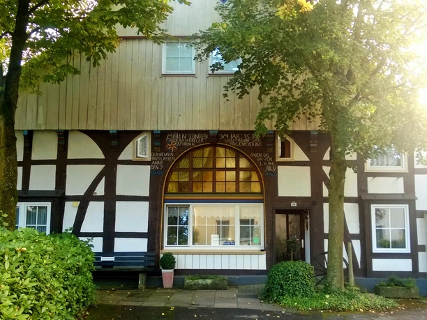 Fachwerkhaus mit heller Holzverkleidung und Bogenfenster. Umgeben von Bäumen und einem Gehweg.