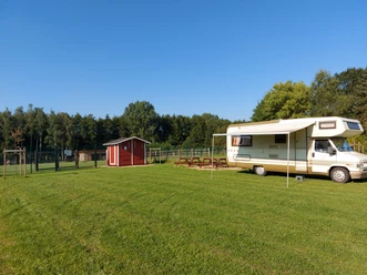 Wohnmobil auf grünem Stellplatz, daneben eine rote Holzhütte; Bäume im Hintergrund bei klarem Himmel.