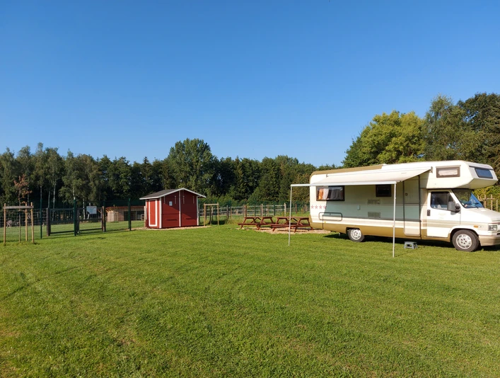 Wohnmobil auf grünem Stellplatz, daneben eine rote Holzhütte; Bäume im Hintergrund bei klarem Himmel.
