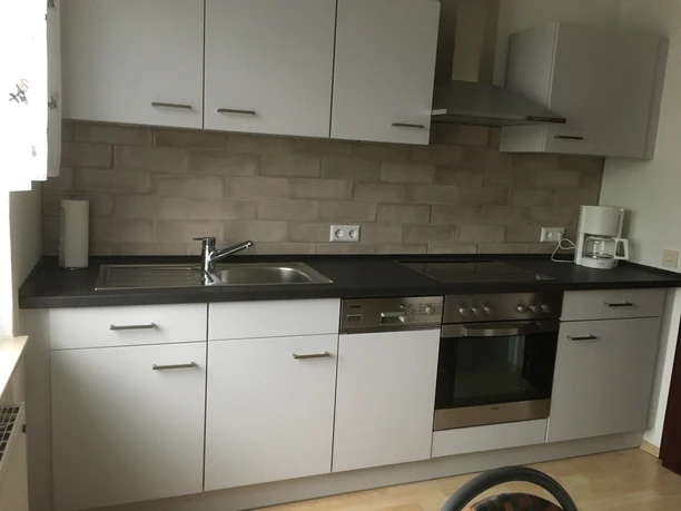 Ferienwohnung Ruhepol Moderne Küchenzeile mit Edelstahlspüle und Elektroherd, umgeben von hellen Holzschränken und Kaffeepadmaschine.