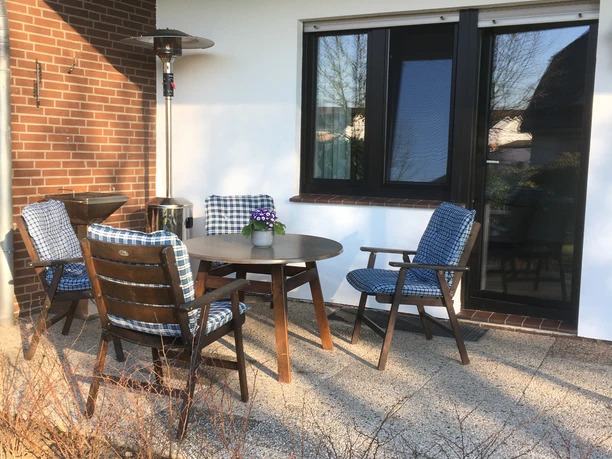 Ferienwohnung Ruhepol Terrasse mit Holzmöbeln und Tisch vor einer weiß getünchten Fassade, umgeben von Heckenpflanzen.