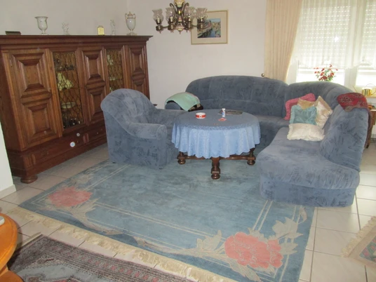 Finke 2.JPG Wohnzimmer mit blauen Polstermöbeln, Holzschrank, Teppich mit Blumenmuster, hellem Boden und Dekor.