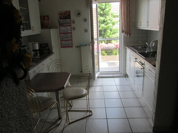 Helle Küche mit weißen Schränken, Esstisch, Stühlen, einer Tür zum Balkon und Blick ins Freie.