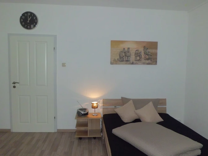 Ein schlicht eingerichtetes Schlafzimmer mit einem Bett, einem Nachttisch, einer Lampe und einer Wanduhr.