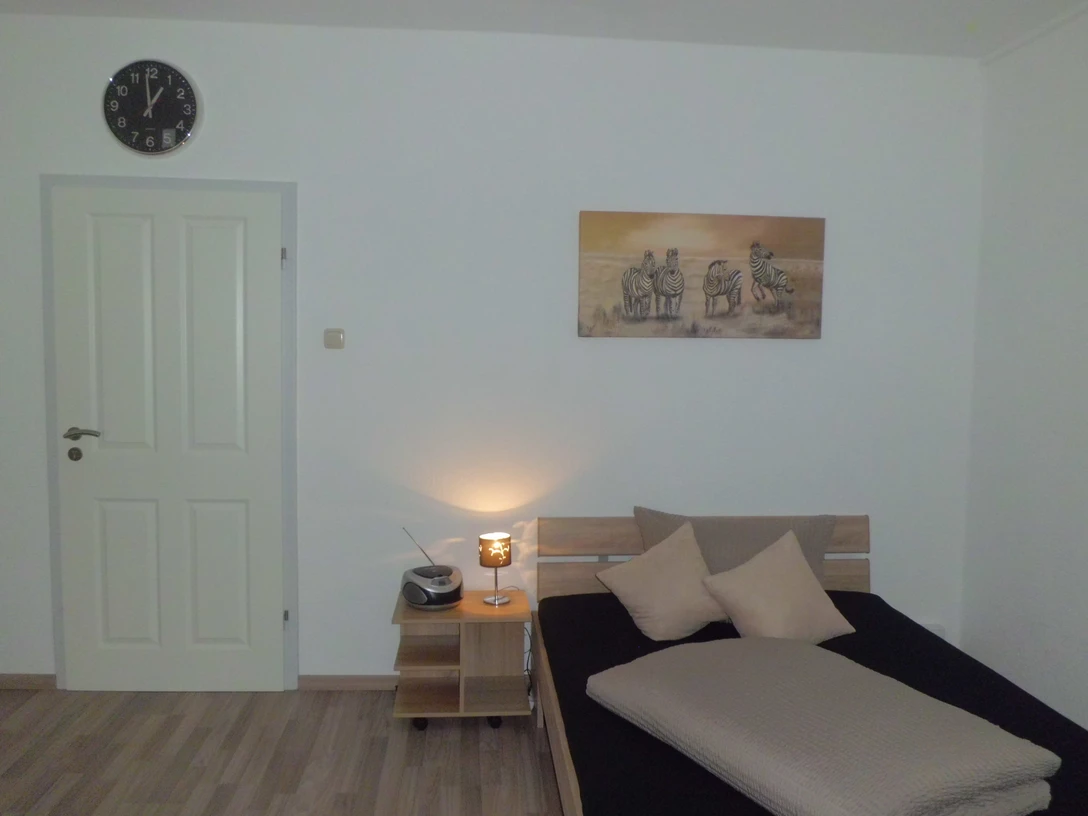 Juritsch.JPG Ein schlicht eingerichtetes Schlafzimmer mit einem Bett, einem Nachttisch, einer Lampe und einer Wanduhr.