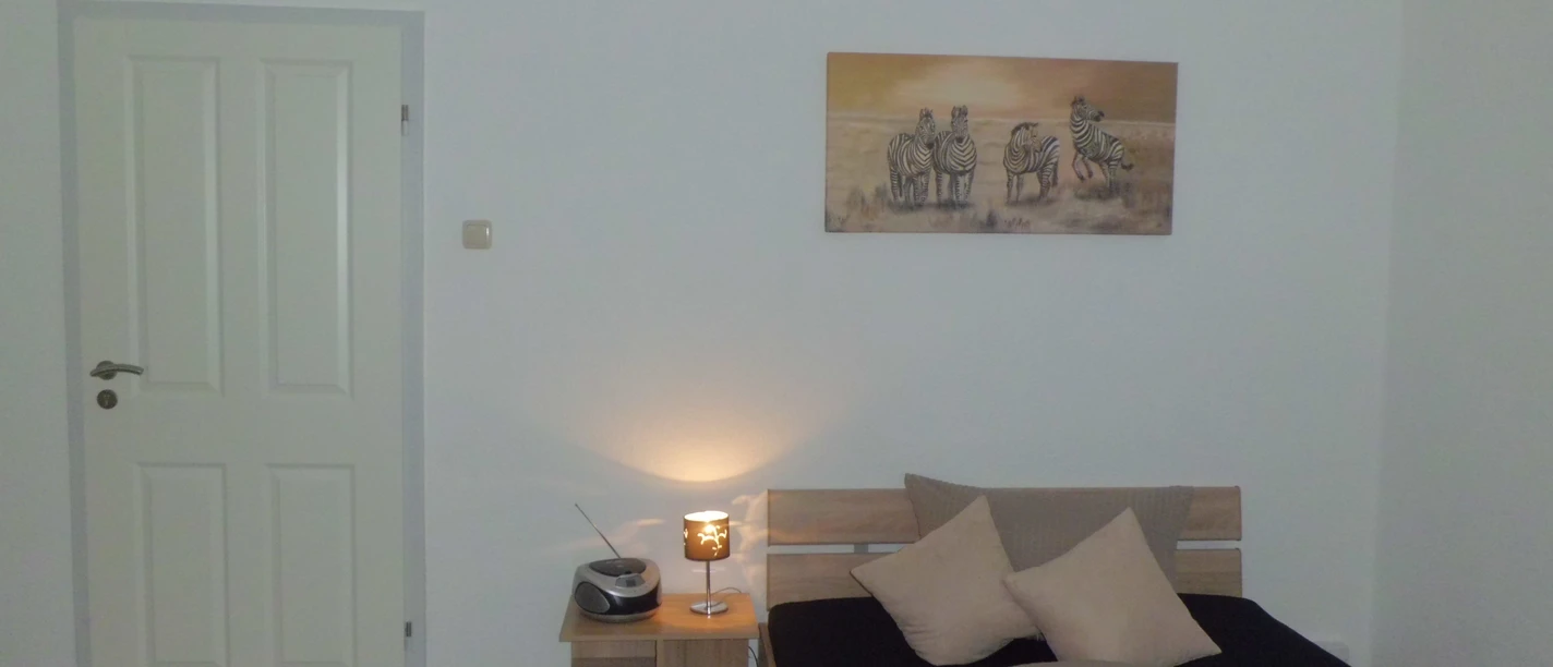 Juritsch.JPG Ein schlicht eingerichtetes Schlafzimmer mit einem Bett, einem Nachttisch, einer Lampe und einer Wanduhr.