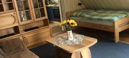 Ein kombinierter Wohn- und Schlafbereich mit Holzmöbeln, Blumen auf dem Tisch und einem Einzelbett.