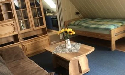 Schlaf_-_und_Wohnraum Dräger.jpeg Ein kombinierter Wohn- und Schlafbereich mit Holzmöbeln, Blumen auf dem Tisch und einem Einzelbett.