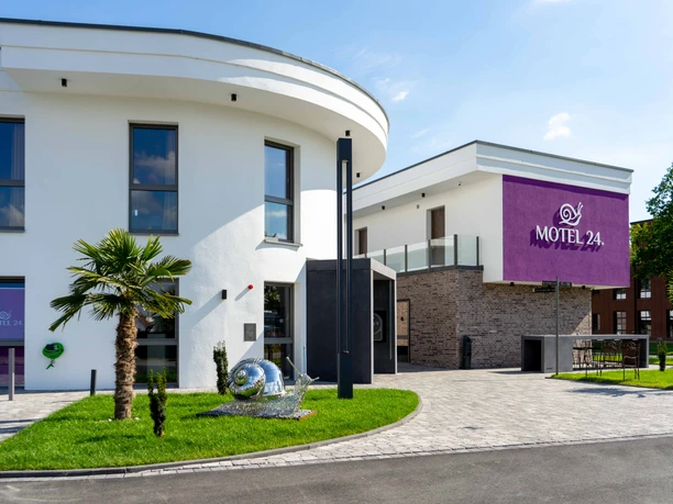 Modernes Motelgebäude mit weißer Fassade und violettem Motel 24h-Logo, umgeben von Rasenflächen.